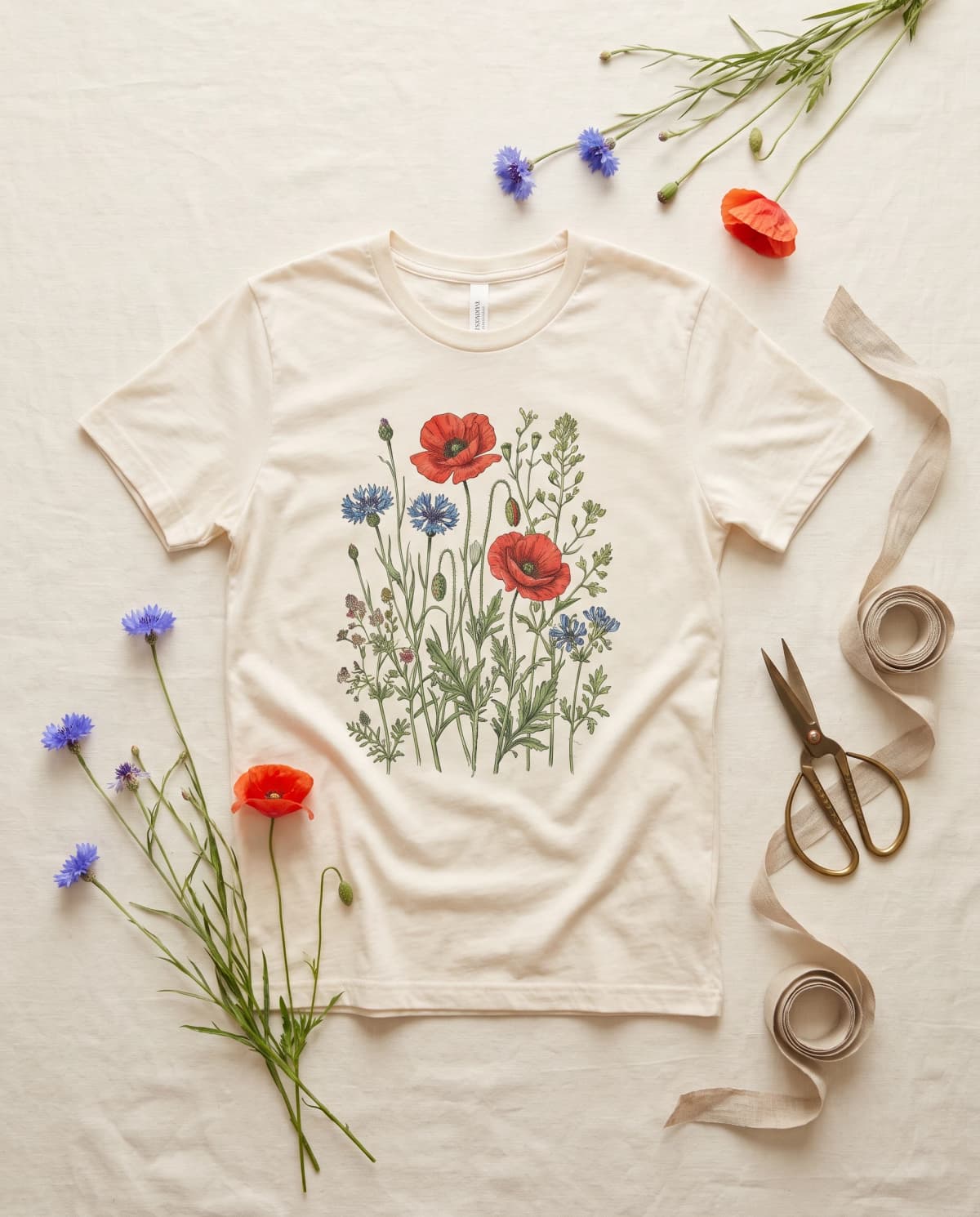 Cremefarbenes T-Shirt mit botanischem Mohn-und-Kornblumen-Print, daneben frische Blumen und Schere