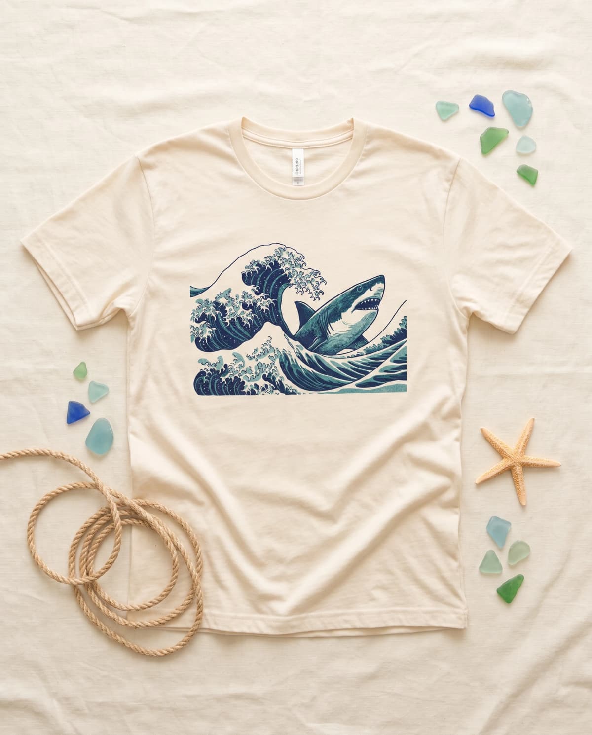 Cremefarbenes T-Shirt mit Hai-und-Welle-Print im Holzschnitt-Stil, daneben Seil, Seeglas und Seestern