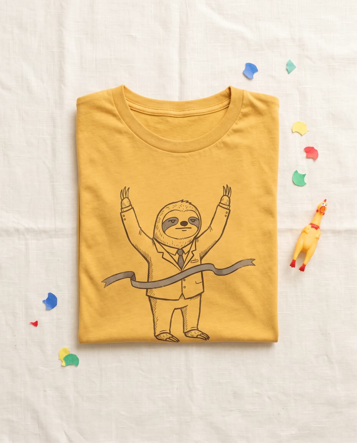 Senfgelbes T-Shirt mit Faultier-im-Anzug-Print, daneben Konfetti und Gummihuhn